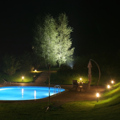 Image d'une piscine éclairée la nuit avec un arbre en arrière-plan. La piscine est rectangulaire et entourée d'une terrasse en bois. L'eau de la piscine est bleue et calme. L'éclairage est doux et crée une atmosphère relaxante. 