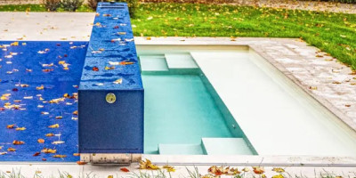 Une piscine extérieure rectangulaire, aux parois blanches, entourée d'une terrasse en pierre claire et d'une pelouse verte. Des feuilles d'automne jonchent la terrasse et la pelouse